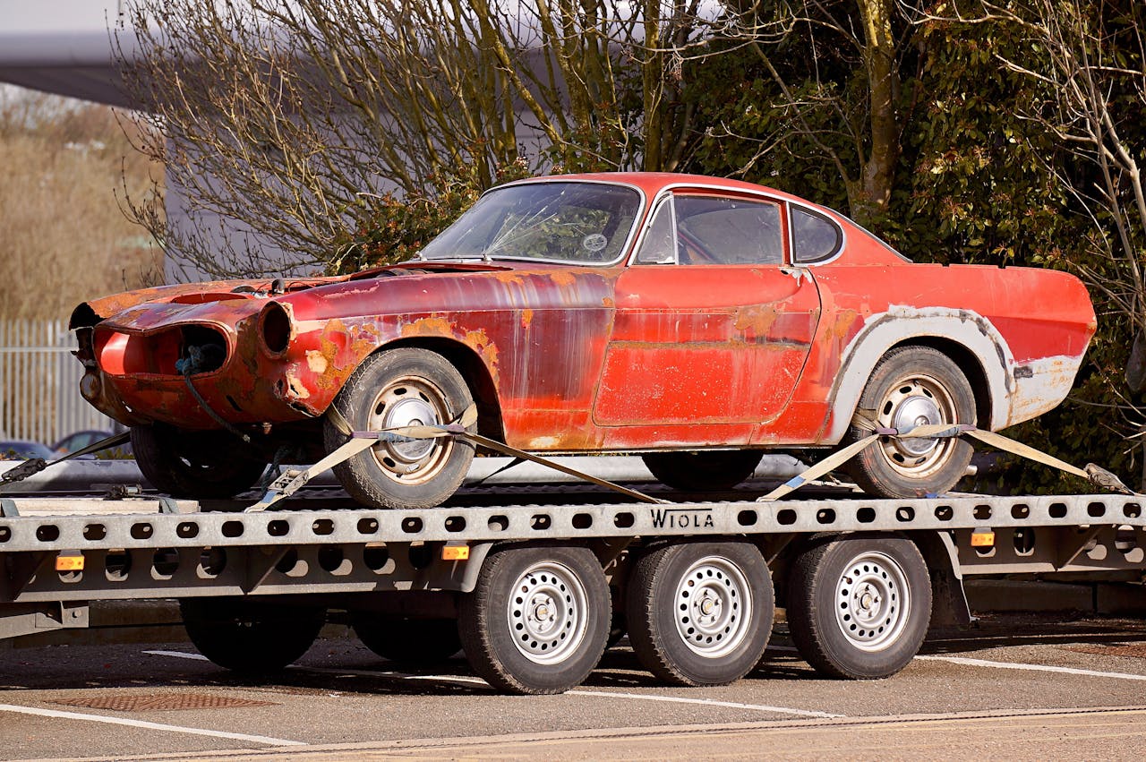 programs-02 A worn-out, rusty vintage car secured on a flatbed trailer, outdoors under daylight.